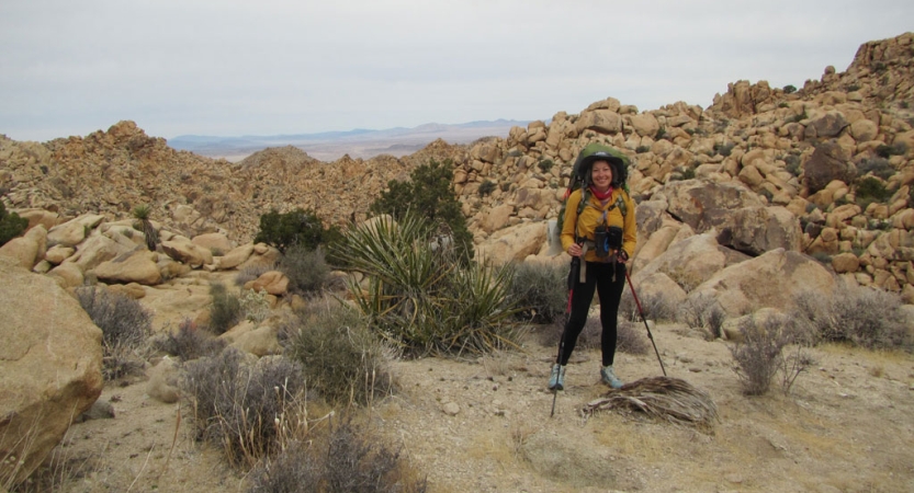 a veteran wearing a large backpacking holds a trekking pole while they stand in front of a rocky landscape in joshua tree